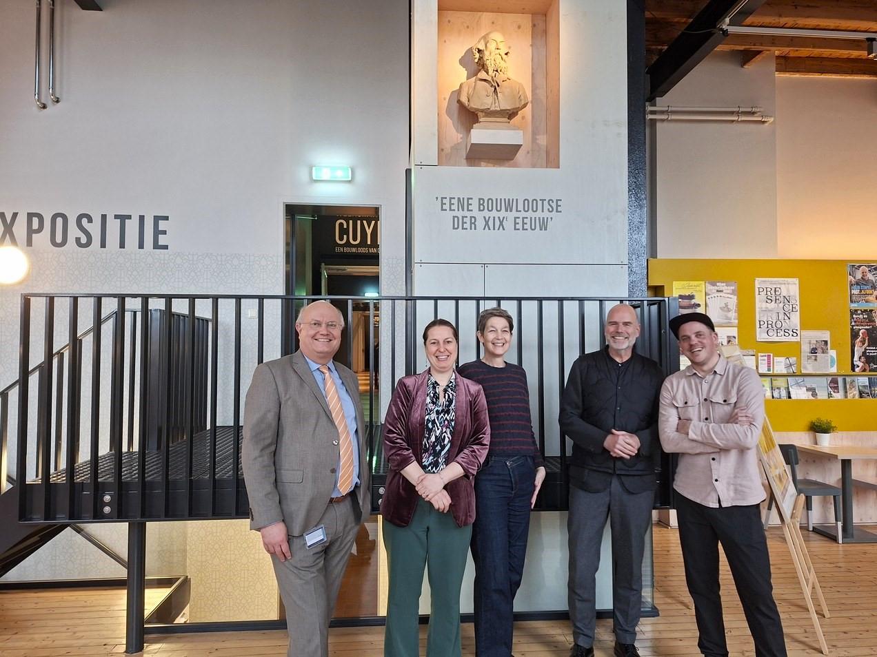 Groepsfoto van vijf lachende personen die de start van de voorbereiding van de terugkeer van de Koninklijke Troon vieren in de hal van het Cuypershuis in Roermond, onder een buste van architect Pierre Cuypers.