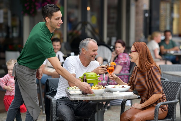 Ober brengt de lunch naar het tafeltje waar een oudere man en vrouw aan het proosten zijn