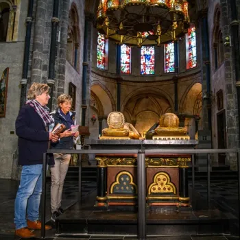 Twee bezoekers staan bij het praalgraf van graaf Gerard III van Gelre en Margaretha van Brabant in de Munsterkerk in Roermond, omringd door romaanse bogen en glas-in-loodramen.