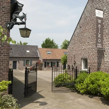 Binnenplaats van d’Ouffenhoff met open sierhek, bakstenen boerderijgebouwen en een welkomstbord, omgeven door groen en rust.